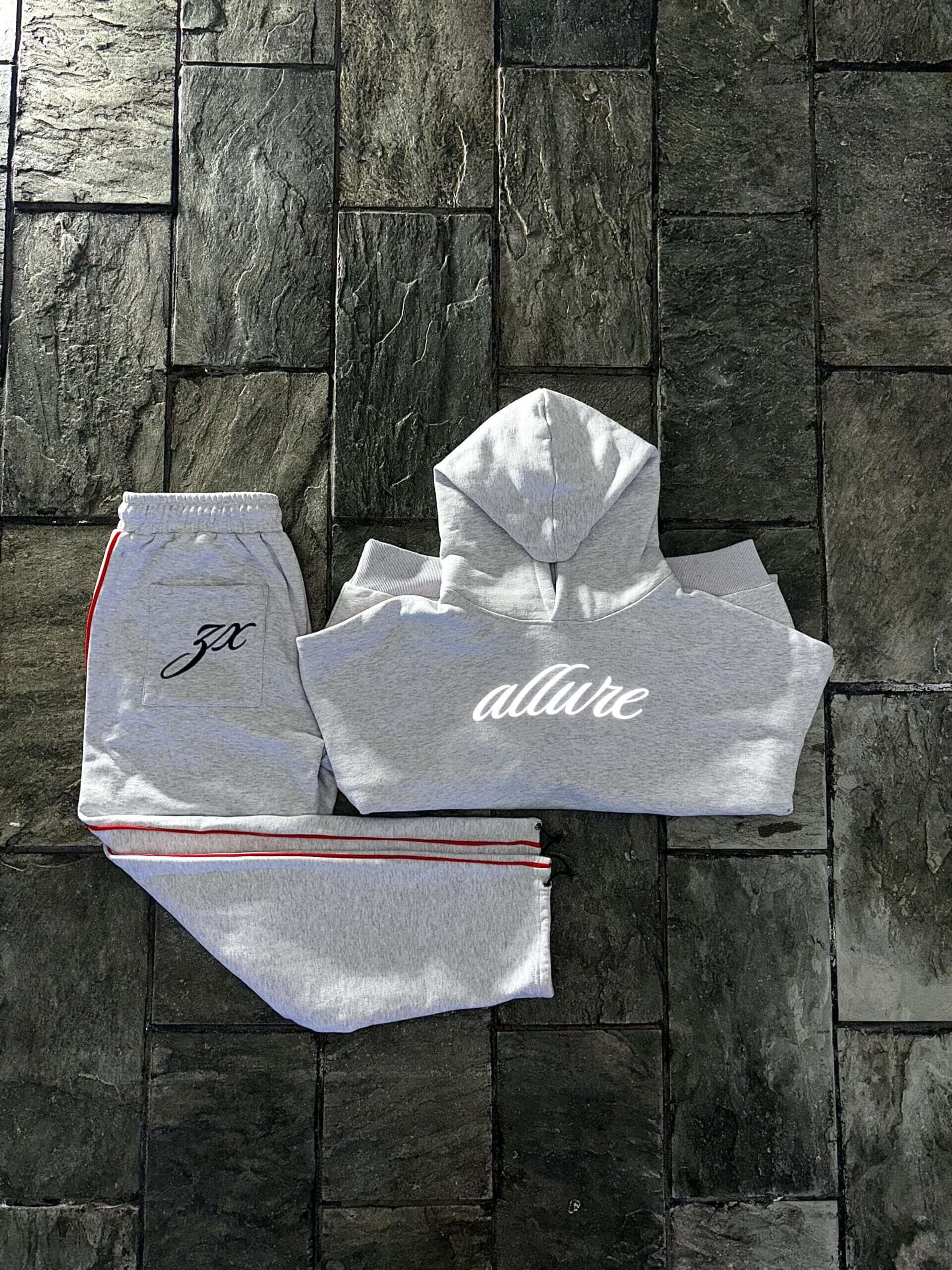 Allure track pants and hoodie in grey with front embroidery and red side stripes on a stone floor.