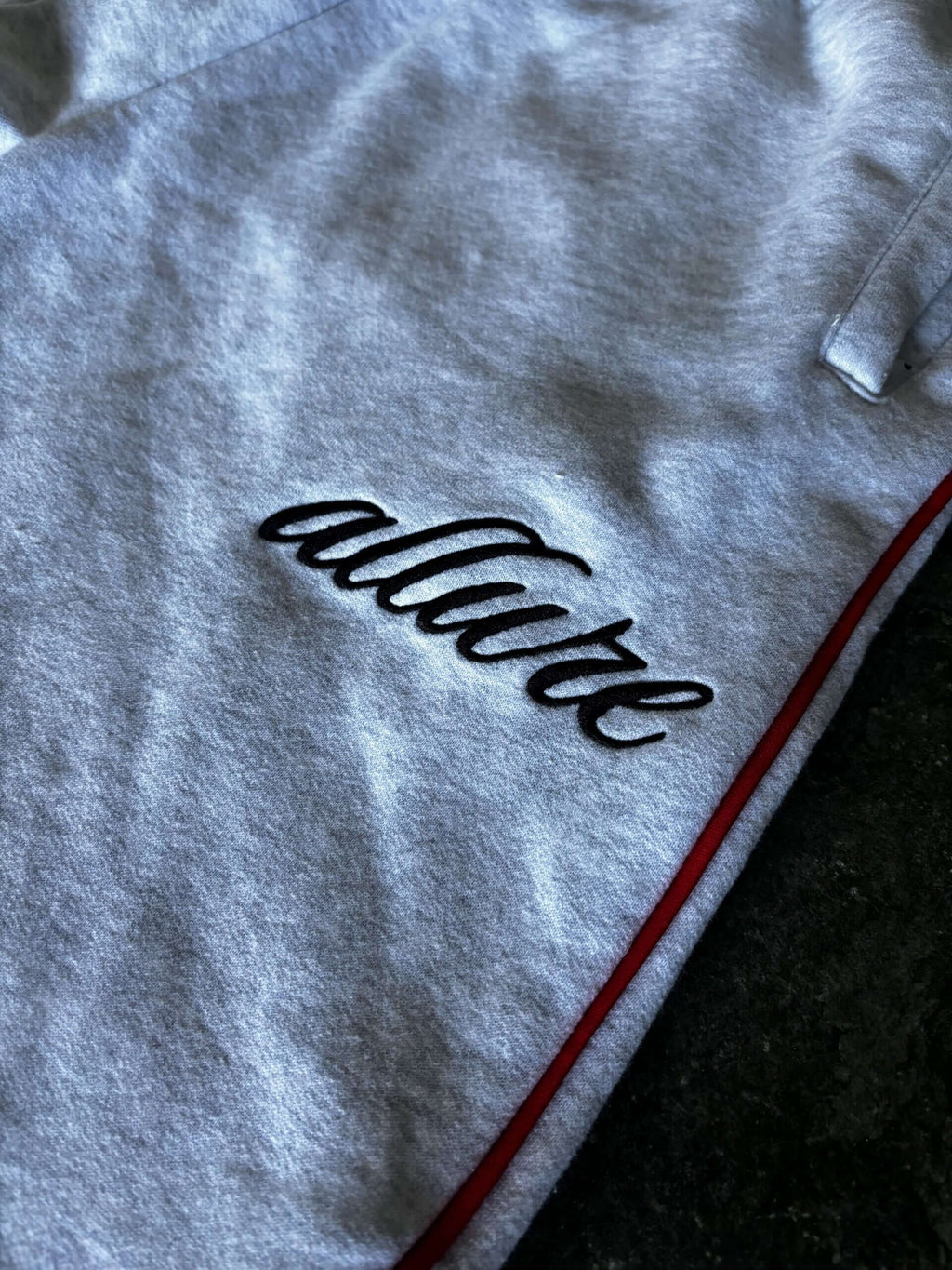 Close-up of Allure Track Pants showing embroidered logo on grey fabric with red stripe accents.