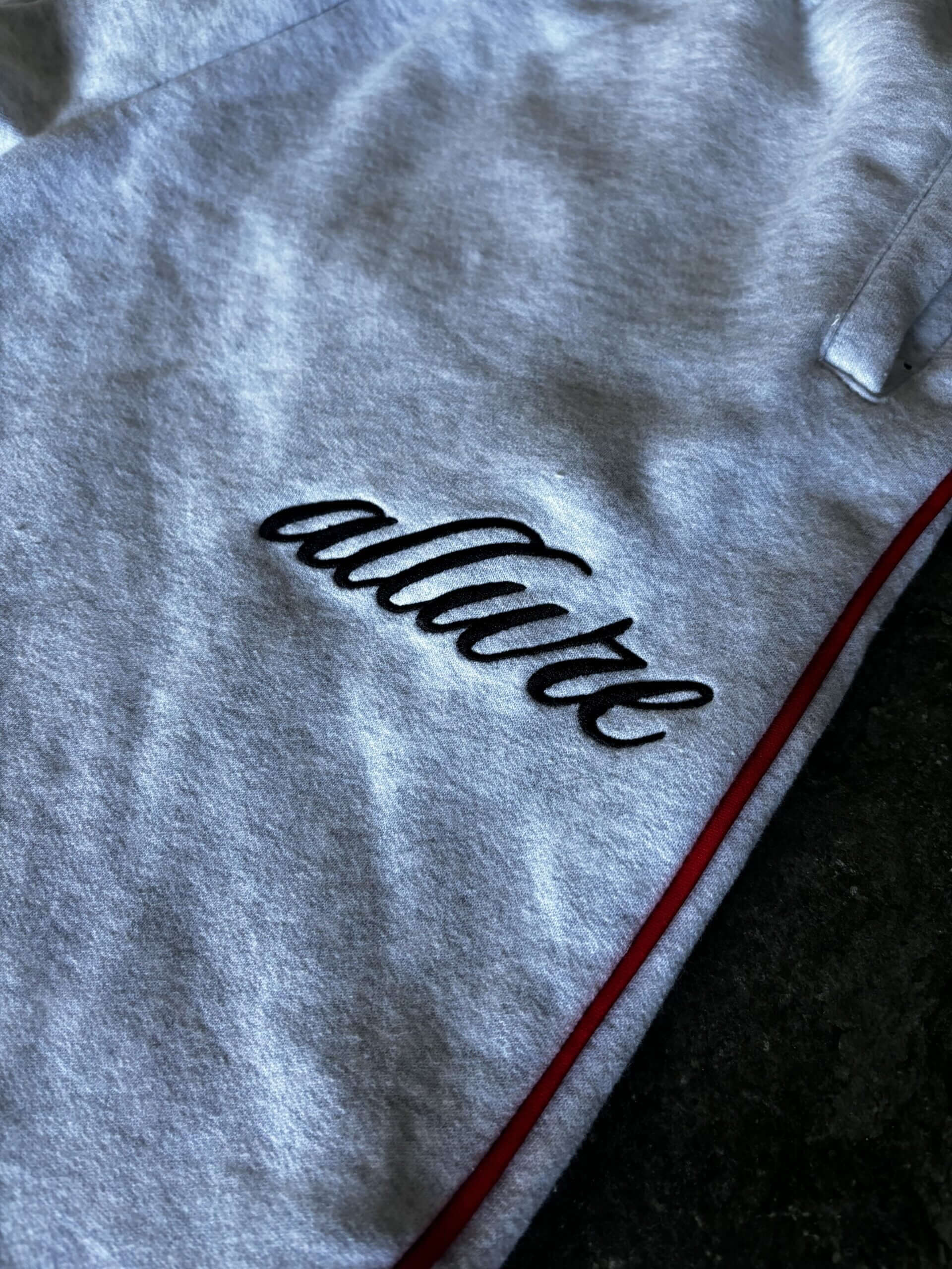 Close-up of Allure Track Pants showing embroidered logo on grey fabric with red stripe accents.